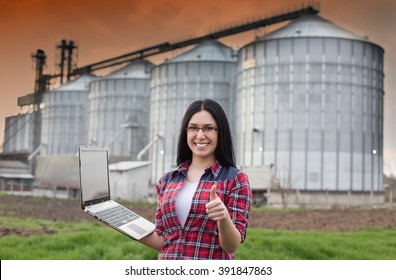 Young smiling farmer girl standing with laptop in front of grain silo and showing ok hand sign