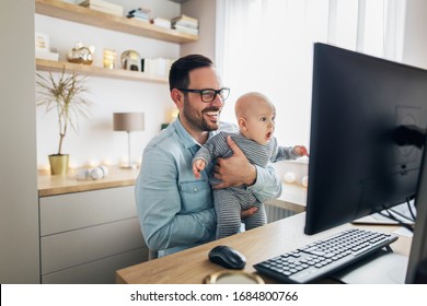 Young smiling father working on computer from home while holding his baby boy in his arms.