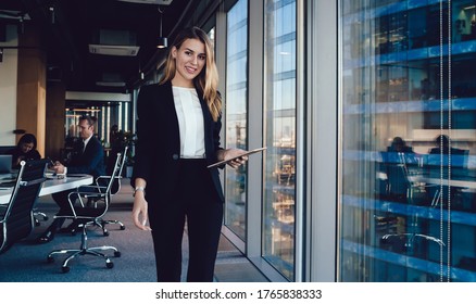 Young smiling female entrepreneur in elegant clothes browsing tablet while standing in  contemporary workplace on background of group of busy colleagues and looking at camera - Powered by Shutterstock