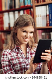 Young Smiling Female Merchant With IPad Tablet In Book Store Or Library. Happy Successful Commerce Woman Over Books Shelf Background.