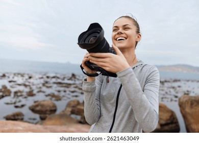 Young smiling female photographer holding a camera outdoors, enjoying nature by the rocky shore, capturing beautiful moments in a vibrant coastal setting