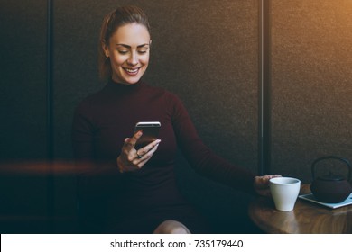 Young smiling female reading text message on mobile phone while resting in coffee shop interior, attractive woman watching funny video on cell telephone while enjoying cup of tea in modern restaurant