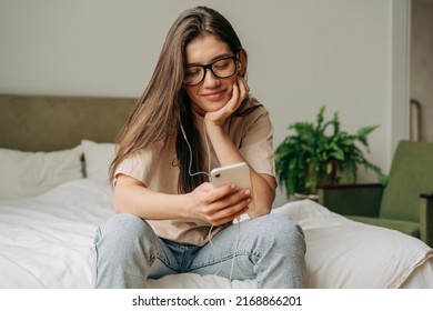 Young Smiling Female Student Holding Cellphone And Scrolling Social Media Feed And Chatting At Home