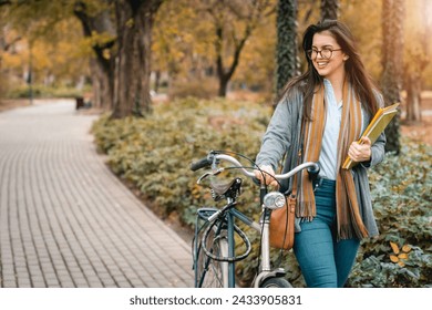 Young smiling female student holding books while walking with bike in public park. Beautiful girl posing with bike outdoors. - Powered by Shutterstock