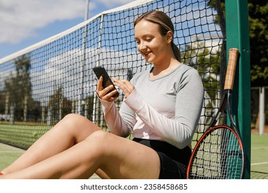 Young smiling female tennis player sitting on court and using smart phone during a break in training - Powered by Shutterstock