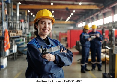 Young smiling female worker of modern industrial plant or factory in workwear and protective helmet standing in large workshop