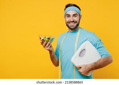 Young smiling fitness trainer instructor sporty man sportsman in headband blue t-shirt measure tape hold plate with vitamin salad hug scales isolated on plain yellow background. Workout sport concept - Shutterstock ID 2138588865