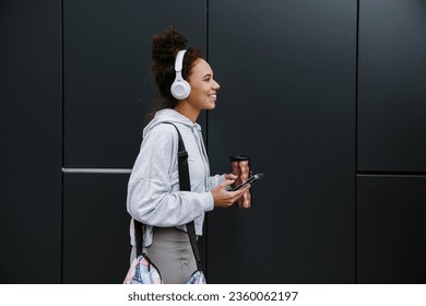 Young smiling fitness woman in sportswear listening music with headphones and mobile phone while standing outdoors near black wall - Powered by Shutterstock