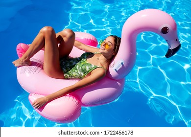 Young smiling fitted girl in bikini relax chilling on pink inflatable flamingo in swimming pool. Attractive woman in swimsuit lies in a sun on tropical vacation. Pretty female sunbathing at resort.