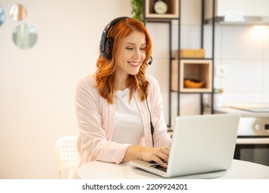 Young Smiling Friendly Ginger Girl Working As Agent At Call Center From Home. Wearing Headset And Typing On Laptop.