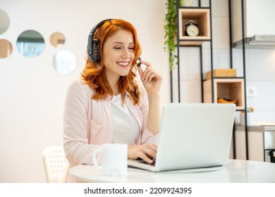 Young smiling friendly ginger girl working as agent at call center from home while drinking coffee. Wearing headset and typing on laptop.