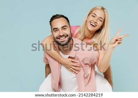 Young smiling fun couple two friends family man woman in casual clothes giving piggyback ride to joyful, sit on back show v-sign gesture together isolated on pastel plain light blue background studio