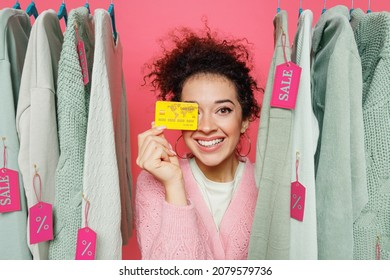 Young Smiling Fun Female Costumer Woman 20s Wear Sweater Stand Near Clothes Rack With Tag Sale In Store Showroom Hold Cover Eye With Credit Bank Card Isolated On Plain Pink Background Studio Portrait