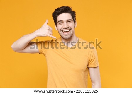 Young smiling fun friendly unshaved caucasian handsome man 20s wears casual basic blank print design t-shirt doing phone finger gesture like call me back isolated on yellow background studio portrait