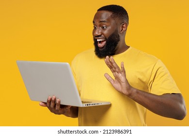Young Smiling Fun Happy Black Man 20s Wearing Bright Casual T-shirt Hold Use Work On Laptop Pc Computer Waving Hand Isolated On Plain Yellow Color Background Studio Portrait. People Lifestyle Concept