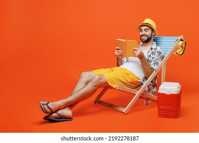 Young smiling fun happy cool tourist man in beach shirt hat lie on deckchair near fridge read book novel isolated on plain orange background studio portrait. Summer vacation sea rest sun tan concept - Powered by Shutterstock