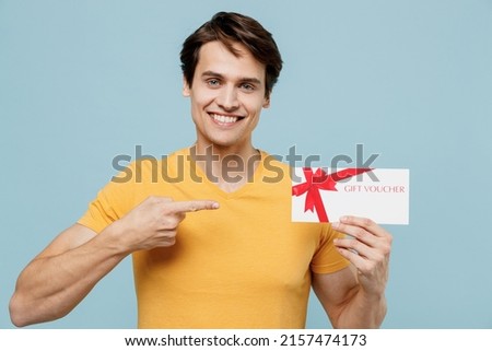 Young smiling fun happy satisfied man 20s wear yellow t-shirt hold point index finger on gift certificate coupon voucher card for store isolated on plain pastel light blue background studio portrait.