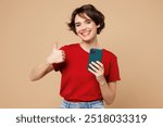 Young smiling fun happy woman she wear red t-shirt casual clothes hold in hand use mobile cell phone show thumb up isolated on plain pastel light beige background studio portrait. Lifestyle concept