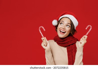 Young smiling fun happy woman 20s wear Santa Claus Christmas red hat hold candy cane stick lollipop look aside isolated on plain red background studio portrait. Happy New Year 2022 celebration concept