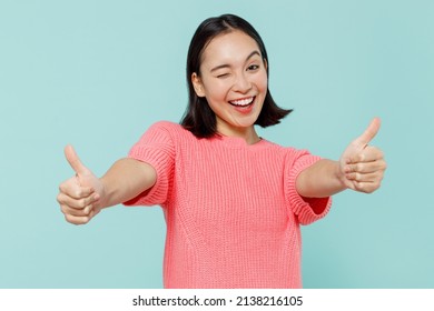 Young Smiling Fun Happy Woman Of Asian Ethnicity 20s Wearing Pink Sweater Showing Thumb Up Like Gesture Blink Isolated On Pastel Plain Light Blue Background Studio Portrait. People Lifestyle Concept