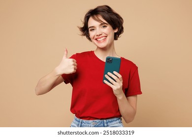 Young smiling fun happy woman she wear red t-shirt casual clothes hold in hand use mobile cell phone show thumb up isolated on plain pastel light beige background studio portrait. Lifestyle concept - Powered by Shutterstock