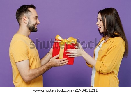 Young smiling fun satisfied impressed couple two friends family man woman together in yellow clothes hold give red present box with gift ribbon bow isolated on plain violet background studio portrait