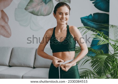 Young smiling fun strong sporty athletic fitness trainer instructor woman wears green tracksuit hold measure tape on waist training do exercises at home gym indoor. Workout sport motivation concept