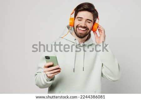 Young smiling fun vivid caucasian man wear mint hoody headphones listen to music dance use mobile cell phone have fun isolated on plain solid white background studio portrait. People lifestyle concept
