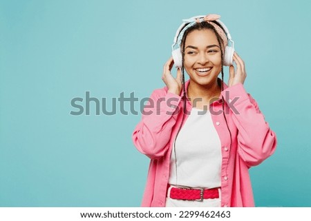 Young smiling fun woman of African American ethnicity wear pink shirt white t-shirt headscarf headphones listen to music look aside isolated on plain pastel light blue cyan background studio portrait