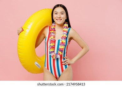 Young smiling fun woman of Asian ethnicity in striped one-piece swimsuit hawaii lei hold inflatable ring stand akimbo isolated on plain pastel pink background. Summer vacation sea rest sun tan concept
