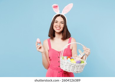 Young smiling fun woman wears pink casual clothes rabbit bunny ears wicker basket hold in hand egg look camera isolated on plain blue cyan background studio portrait. Lifestyle Happy Easter concept