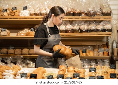 Young smiling girl with bread in the bakery, preparing pastry for sale in supermarket bakery department. Bakery seller.
