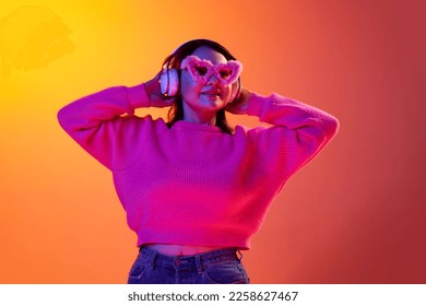 Young smiling girl in cozy sweater  fur glasses listening to music in headphone over gradient orange background in neon light  Concept emotions  facial expression  youth  inspiration  sales  ad