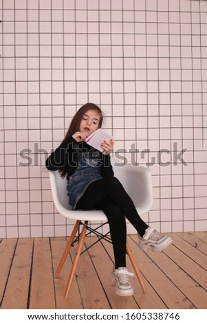 young smiling girl of eight years with long hair in a black turtleneck and jeans sundress sits with a notebook grimaces