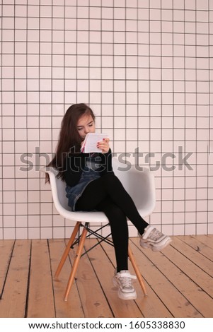 young smiling girl of eight years with long hair in a black turtleneck and jeans sundress sits with a notebook grimaces