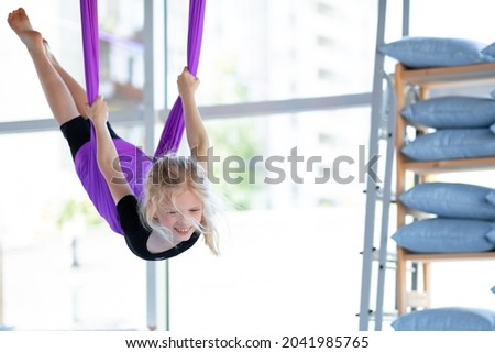  young smiling girl practice in aero stretching swing in purple hammock in fitness club. kids Aerial flying yoga exercises.