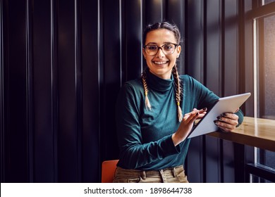 Young Smiling Girl Sitting In Cafe Near Window, Scrolling On Tablet And Looking At Camera.