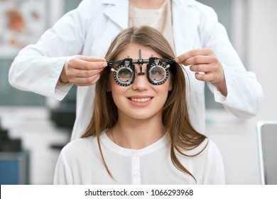 Young Smiling Girl Is Sitting And Looking At Camera Through During Vision Checking. Ophthalmologist Is Using Special Medical Equipment For Eye Health Saving And Improving.