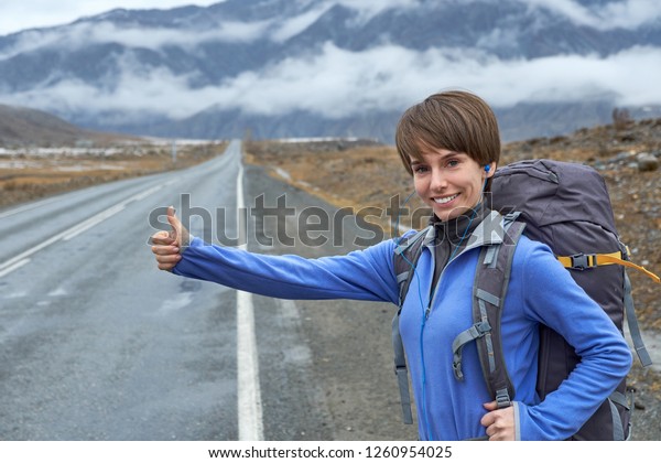 A young smiling\
girl is traveling in the mountains. stops the car on the road\
(hitchhiking), raises his\
hand
