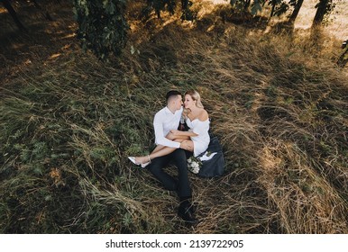 A young, smiling groom and a cute blonde bride with curly hair in a white dress are sitting on a rug on the yellow grass in the fall in nature and hugging. Wedding photo of the newlyweds, top view. - Powered by Shutterstock