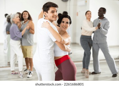 Young smiling guy learning to dance waltz paired with expressive positive Hispanic girl in dancing class