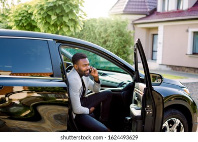 Young smiling handsome African businessman sitting in the car on passenger seat and talking on the phone. Building, house and green trees on teh background