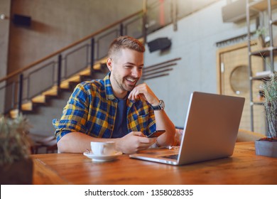 Young smiling handsome Caucasian unshaven freelancer in plaid shirt sitting at restaurant and using smart phone. In front of him laptop. - Powered by Shutterstock