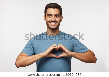 Young smiling handsome smiling male in blue t-shirt showing heart sign isolated on gray background