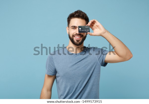 Young smiling handsome man in casual clothes posing isolated on blue wall background studio portrait. People sincere emotions lifestyle concept. Mock up copy space. Covering eye with credit bank card