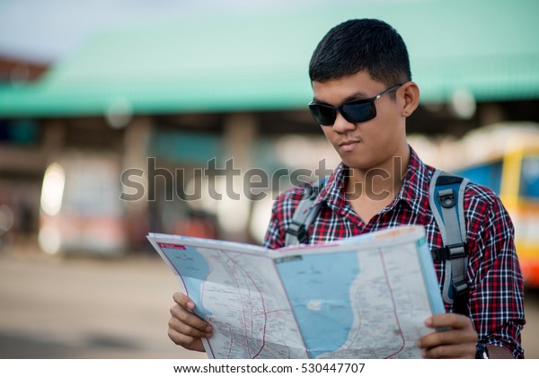 Young smiling handsome man with luggage bag on rainy\
city street with busy traffic transport looking map, waiting for\
public bus