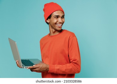 Young Smiling Happy African American Man In Orange Shirt Hat Hold Use Work On Laptop Pc Computer Look Aside Isolated On Plain Pastel Light Blue Background Studio Portrait. People Lifestyle Concept.