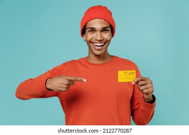 Young Smiling Happy African American Man In Orange Shirt Hat Hold In Hand Point Index Finger On Credit Bank Card Isolated On Plain Pastel Light Blue Background Studio Portrait People Lifestyle Concept