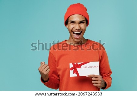 Young smiling happy african american man 20s wear orange shirt hat hold gift certificate coupon voucher card for store do winner gesture celebrate isolated on plain pastel light blue background studio
