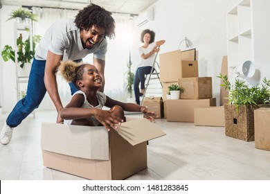 Young Smiling Happy African-american Family Unpacking During Move. New Home. Little preschooler daughter sitting in cardboard box, father rolling her. Smiling mother looking them. - Powered by Shutterstock